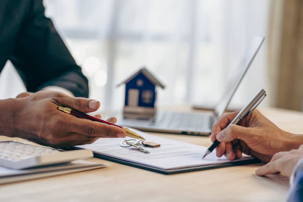 Deux personnes signent un contrat immobilier sur une table. Une main pointe le document tandis que l'autre signe, avec une maison miniature et des clés.